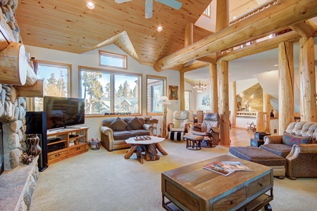 A spacious wooden living room with high ceilings, large windows, various seating arrangements, a stone fireplace, flat-screen TV, and wooden furniture. Light streams in through multiple windows.