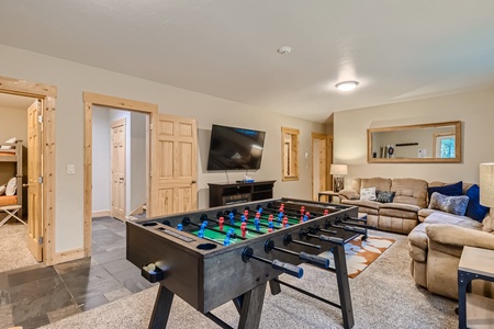 A game room with a foosball table, a large wall-mounted television, a sectional sofa, a coffee table, and a mirror. There are doorways leading to other rooms.