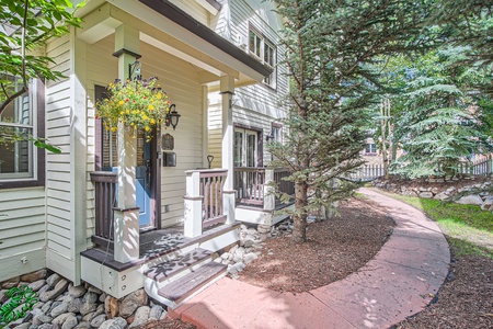 A white and gray house with a front porch featuring hanging plants and a black railing. A curved paved pathway is surrounded by trees and landscaped ground.