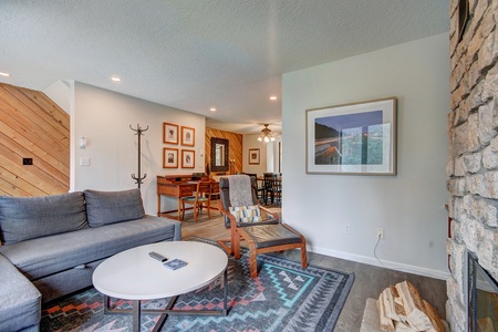 A living room with a gray couch, a wooden chair, a round white coffee table, and a framed picture on the wall. There is a rug on the floor and a wooden wall panel on the left.