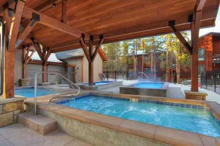 Outdoor hot tub area with multiple heated pools and wooden pergola, surrounded by trees and buildings.