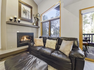 Living room with a leather sofa, cushions, a coffee table, and a lit fireplace on the side. Large windows and a glass door provide views of trees outside.