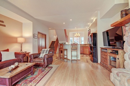 A cozy living room with wooden floors, a stone fireplace, comfortable seating, and a view of the adjacent dining area and kitchen in the background.