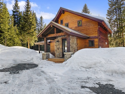 A two-story wooden cabin with a stone porch is surrounded by snow and pine trees under a clear blue sky.