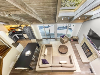 Overhead view of a modern living room with an L-shaped couch, a central round coffee table, a dining area, an open kitchen, and a large window with a snowy scene outside. Exposed wooden ceiling beams.