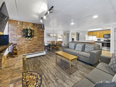 Spacious living room with two gray sofas, wooden coffee table, brick fireplace, and a view of the dining area and kitchen in the background. The room has a wooden floor and white ceiling tiles.