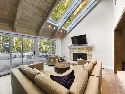 A living room with beige sofas, a circular coffee table, a fireplace with a stone facade and TV above it, and large windows offering a snowy forest view. Skylights are set into the wooden ceiling.