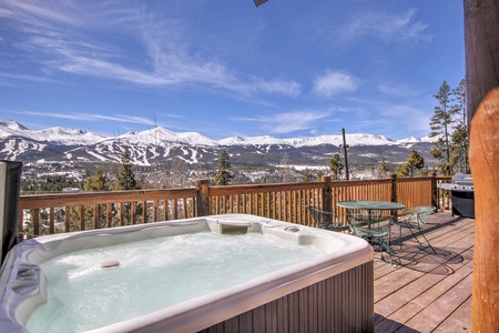 A bubbling hot tub sits on a wooden deck overlooking a scenic view of snow-capped mountains and a ski resort. There is also a metal table with four chairs on the deck.