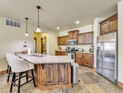 Modern kitchen with wooden cabinetry, stainless steel appliances, granite countertops, and a central island with bar stools. Pendant lights hang above the island, and a pantry is visible in the background.