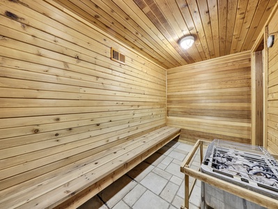 A wooden sauna room with a long bench along the wall and a heater with stones in the foreground under a ceiling light.