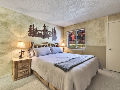 A bedroom with a rustic theme, featuring a double bed with grey bedding, wooden side tables, lamps, and a metal tree wall art. A window with blinds allows natural light to illuminate the room.