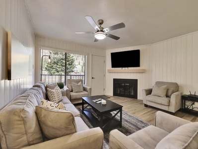 View of the newly updated, light, and airy living room featuring a 58" smart TV, electric fireplace, ceiling fan, and comfortable sofa and armchairs. A glass door leads to the private walk-out balcony