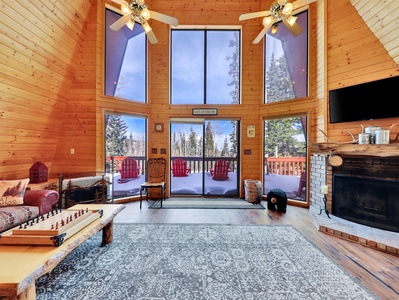 Great room featuring expansive tree views, access to an outdoor deck with a seating area, a cozy wood-burning fireplace, ceiling fans for added comfort, board games, a 43" Smart TV, and plush, comfortable seating.