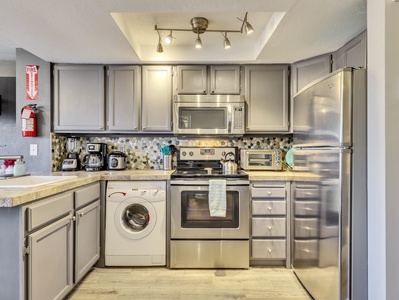 A modern kitchen with stainless steel appliances, grey cabinets, mosaic tile backsplash, washing machine, fire extinguisher, and track lighting.