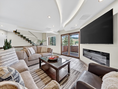 Cozy living room with a flat-screen TV, gas fireplace, and floor-to-ceiling windows opening to a balcony with views of the pond and Bristlecone Park