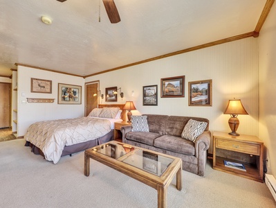 A cozy bedroom with a bed, a brown sofa, a glass-top coffee table, and a nightstand with a lamp. The room has beige walls adorned with framed pictures and a ceiling fan above.