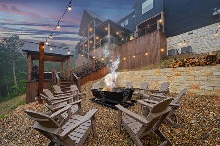 Outdoor fire pit area with brown Adirondack chairs arranged in a circle, surrounded by string lights and a modern cabin with multiple decks in the background, set against a sunset sky.