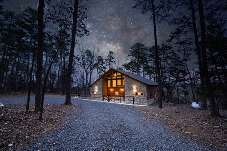 A cabin with exterior lights on, surrounded by trees and a gravel driveway, under a starry sky at night.