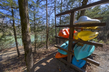 Kayaks in various colors are stored on a wooden rack in a forested area next to a body of water on a sunny day.