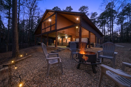 A cozy cabin with a covered porch is surrounded by trees at dusk. In the foreground, there are chairs arranged around a lit fire pit on a gravel surface.