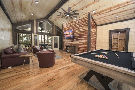 A spacious, wood-paneled game room features a pool table, brown leather furniture, a large flat-screen TV, and windows letting in natural light with a view of trees.