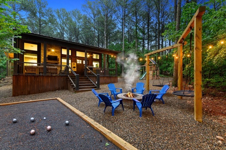 Come ready to enjoy all this cabin ahs to offer.  Surrounded by tall trees, a fire pit with adirondack chairs, swings for the kids, and a bocce ball court are visible. Strings of lights hang above.