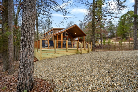 A small wooden cabin with a raised deck and outdoor seating stands among tall trees, surrounded by a gravel area and forest.