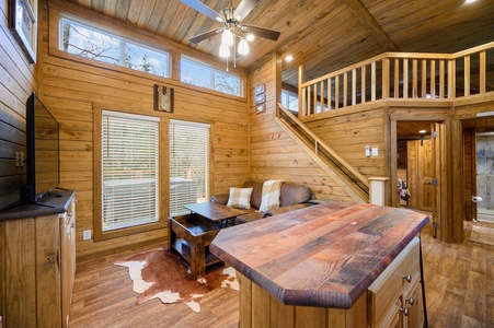 A cozy wooden cabin interior featuring a living area with a couch, coffee table, TV, and loft-style staircase. The space is decorated with a cowhide rug and a ceiling fan with lights.