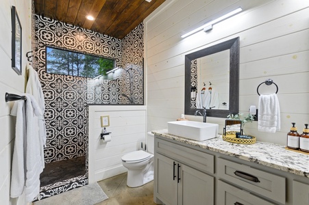 A modern bathroom with patterned tile shower, white shiplap walls, granite countertop with a vessel sink, large mirror, and wooden ceiling. Towels and toiletries are neatly arranged.