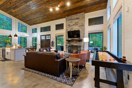 A spacious, modern living room with a high wooden ceiling, a stone fireplace, large windows, and a mix of leather and fabric furniture. You can also try your luck on the shuffleboard game pictured on the right.