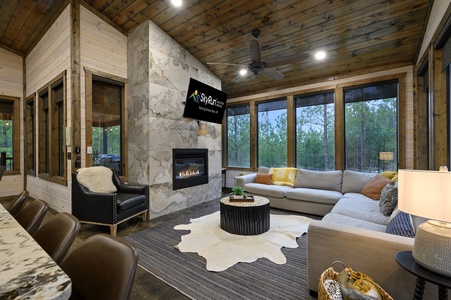 A modern living room with a fireplace, large sectional sofa, and flat-screen TV. The room features wooden walls, large windows, and a cowhide rug.