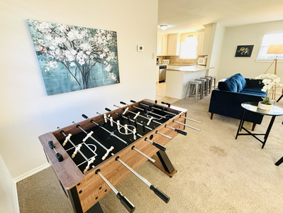 A foosball table in a carpeted room positioned near a window with blinds next to a wall. A painting of a flowering tree hangs above the table. A thermostat is visible on the adjacent wall.