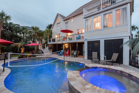 A multi-level house with a patio overlooks a backyard pool and a hot tub, surrounded by lounge chairs and red umbrellas.