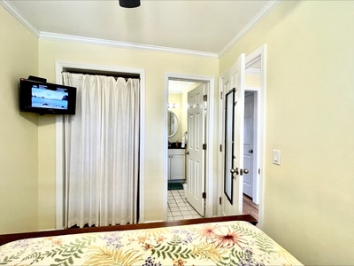 A small bedroom with a floral bedspread, wall-mounted TV, and open doorways leading to a bathroom and closet with a curtain.