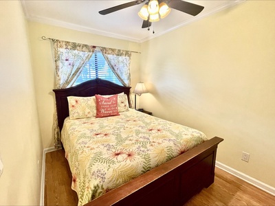 A bedroom with a wooden bed frame, floral bedding, a side lamp, and draped curtains under a ceiling fan.