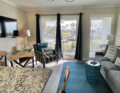 A living room with a striped chair, gray sofa, wall-mounted TV, blue rug, and a balcony view of trees and a road outside large windows.