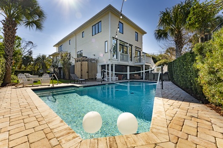 A modern two-story house with a backyard pool, surrounded by patio furniture and palm trees, under a clear blue sky.