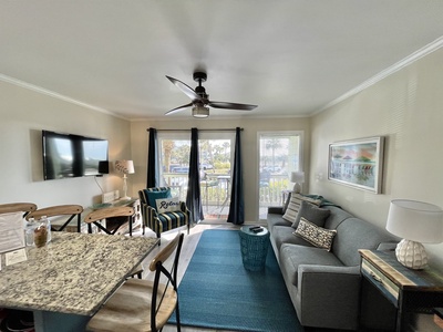 Modern living room with a gray sofa, wall-mounted TV, and ceiling fan. Balcony access through glass doors. Decor includes art and a lamp.