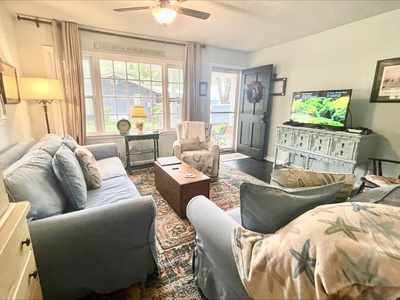 A cozy living room with two blue sofas, a wooden coffee table, and a flat-screen TV on a cabinet. A large window lets in natural light. A fan hangs from the ceiling.