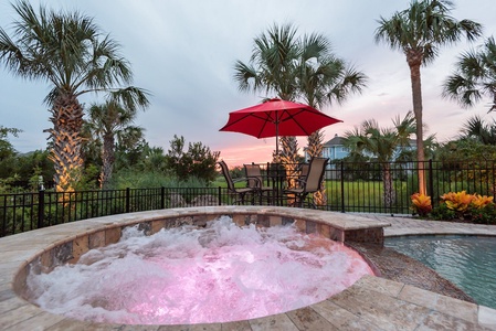 A bubbling hot tub with pink lighting next to a patio set and red umbrella, surrounded by palm trees and a black fence, with a sunset sky in the background.