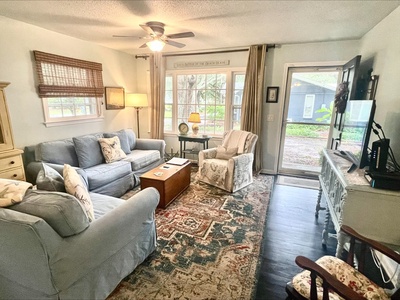 A cozy living room with two sofas, an armchair, a coffee table, a TV on a cabinet, and large windows. Walls are light-colored with various decorations, and a decorative rug covers the dark flooring.