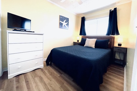 Bedroom with a large bed, dark blue bedding, and two pillows. There's a white dresser with a TV, two nightstands with lamps, and a starfish picture on the wall. Light wood floor.