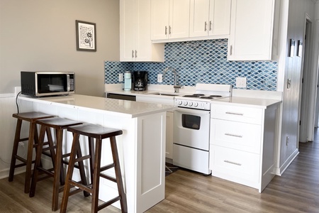 Compact kitchen with white cabinets, a small island with two wooden stools, a microwave, coffee maker, and stove. Blue patterned tile backsplash adds a splash of color.