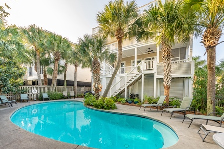 Clear blue kidney shaped pool with chaise lounge chairs, palm trees in a fenced backyard. Stairs rise left to right to the porch overlooking the pool.