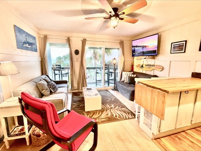 Cozy living room with two sofas, a red chair, ceiling fan, wall art, and large windows overlooking a balcony with outdoor furniture.