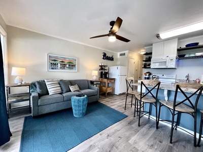 A cozy living room and kitchen area with a gray sofa, blue rug, black bar stools, and various beach-themed decor. A ceiling fan and wall art are visible.