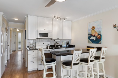 A modern kitchen with white cabinets, black countertops, and four white bar stools. A colorful painting hangs on the wall above the counter. A hallway with a door is visible in the background.