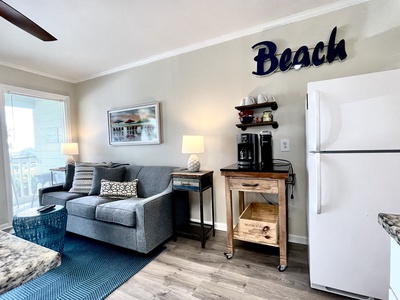 Cozy living room with a gray sofa, blue rug, and coffee table. Wall art and lamps frame the sofa. A wooden cart with a coffee maker is next to a white fridge. "Beach" sign on the wall.