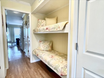 A hallway features bunk beds with floral bedding and beige walls, leading to a living area with wooden flooring and a window view.