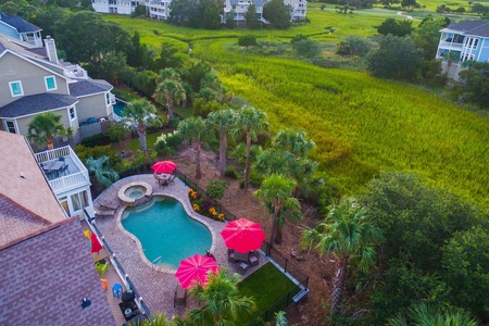 Aerial view of a backyard with a pool, red umbrellas, and surrounding palm trees. Nearby are houses and a grassy field.